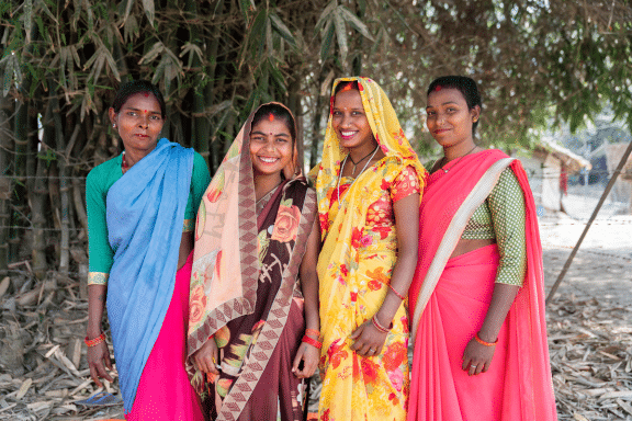 photo of a group of 4 women smiling and wearing colourful saris standing in front of a tree